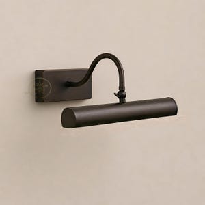 Könnte beinhalten: Eine bronzefarbene Bildleuchte. Die Leuchte hat eine rechteckige Basis, einen gebogenen Arm und ein zylindrisches Leuchtengehäuse. Diese Wandleuchte ist für die Beleuchtung von Kunstwerken oder anderen Objekten konzipiert.