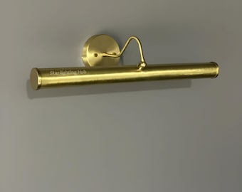 May include: A brass-colored picture light fixture. The fixture has a circular base, a curved arm, and a cylindrical light bar. The text "Star lighting Hub" is printed on the light bar. The light is mounted on a gray wall.