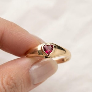 May include: A gold signet ring featuring a single, small, heart-shaped red gemstone set into the center of the band. The ring has a polished, smooth metal finish and a tapered design that widens at the top to hold the vibrant red heart stone.
