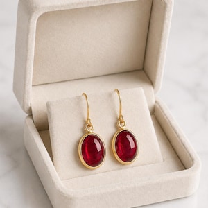 May include: A pair of gold-colored earrings with oval, deep red gemstones, displayed in an open, cream-colored jewelry box. The earrings have a simple, elegant design, with the gemstones set in a gold frame. The jewelry box is lined with a soft, velvety material.