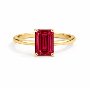 May include: A gold ring featuring a rectangular, deep red gemstone. The gemstone is the focal point, set in a four-prong setting. The band is a simple, polished gold. The ring is displayed against a white background.