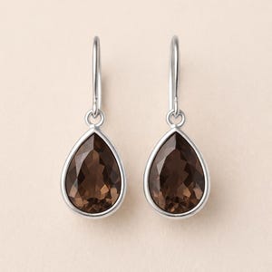 Pode incluir: Um par de brincos pendurados prateados. Cada brinco apresenta uma pedra preciosa castanha facetada em forma de gota, engastada numa moldura prateada. Os brincos penduram-se de um simples gancho prateado.