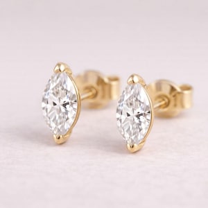 May include: A pair of gold stud earrings with marquise-cut diamonds. The diamonds are set in a bezel setting, and the earrings have a post and butterfly backing. The earrings are displayed on a light pink surface.