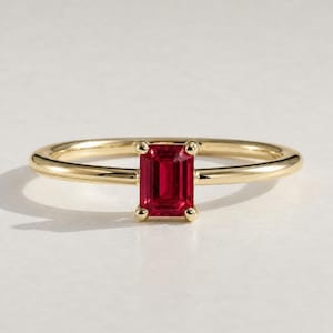 May include: A gold ring featuring a rectangular, deep red gemstone. The ring has a thin, polished band and four small prongs securing the gemstone. The setting and band have a bright, reflective finish.