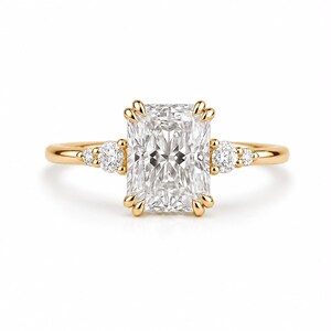 May include: A gold ring featuring a large rectangular radiant-cut center stone held by four prongs. The band is crafted from polished yellow gold and includes two smaller round-cut accent stones on each side of the central setting.