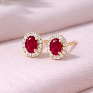 Puede incluir: Un par de pendientes de oro con piedras preciosas rojas oscuras ovaladas rodeadas por un halo de pequeñas piedras preciosas claras. Los pendientes están colocados sobre una tela rosa claro.