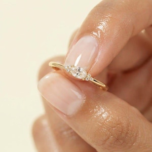 Könnte beinhalten: Ein goldener Ring mit einem zentralen, oval geschliffenen, klaren Edelstein, der auf jeder Seite von drei kleinen, rund geschliffenen, klaren Steinen flankiert wird. Das Band ist dünn und poliert und zeigt ein schlichtes Design mit glänzendem Finish.