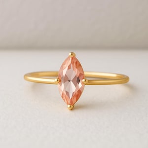 Puede incluir: Un anillo de oro con una gema de color melocotón tallada en forma de marquesa. La gema está engastada con pequeños detalles dorados. El anillo tiene una banda fina y lisa.
