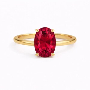 May include: A gold ring featuring a large, oval-cut ruby gemstone. The ruby is a vibrant red color and is held in place by four prongs. The ring's band is a simple, polished gold.