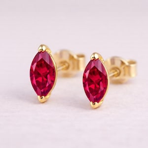 Peut inclure: Une paire de boucles d'oreilles à clous en or avec des pierres précieuses rouges vives en forme de marquise. Les boucles d'oreilles ont une finition dorée polie et sont posées sur un fond blanc cassé doux. Les pierres sont facettées.