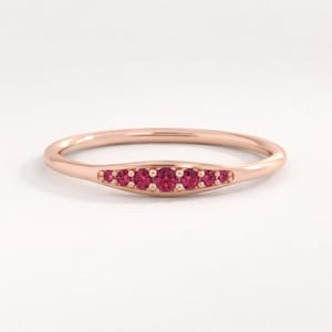 以下が含まれることがあります： 7つの小さな丸い鮮やかな赤い宝石がセットされた、ローズゴールドのリング。リングのデザインはシンプルです。背景はプレーンな白です。