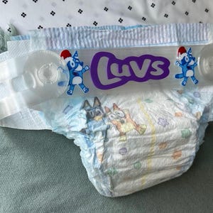 Op de afbeelding: Een wegwerpluier met een lichtblauw en wit gestreepte tailleband en een patroon van getekende honden. Een doorzichtige plastic band is aan de voorzijde bevestigd, met de paarse tekst Luvs en twee blauwe getekende honden met rode mutsjes.