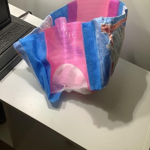 Pode incluir: Uma peça de vestuário absorvente descartável com um painel central de plástico rosa brilhante translúcido, painéis laterais azuis com um padrão texturizado e um forro interior branco. O exterior apresenta uma impressão gráfica parcial com elementos de texto em laranja e branco.