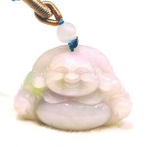 May include: A carved light lavender and pale green jadeite pendant depicting a smiling Buddha figure. The pendant is suspended from a blue cord with a small matching jade bead accent at the top, featuring a smooth, polished, and translucent surface.
