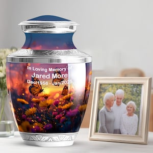 May include: A decorative urn with a sunset and butterfly design. The urn has a silver base and lid, with the text "In Loving Memory of Jared More Dec/1956 - Jan/2025". A framed photo of three people is next to the urn.