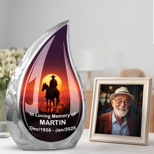 May include: A silver teardrop-shaped memorial urn with a sunset scene of a cowboy on horseback. The urn has the text "In Loving Memory of MARTIN Dec/1956 - Jan/2025". A framed portrait of an older man is next to the urn.