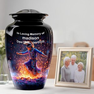 May include: A black cremation urn featuring a glossy finish and a lid. The front displays the text In Loving Memory of madison Dec/1956 - Jan/2025 above a colourful graphic of a person dancing near a fire with bright orange and purple sparks.