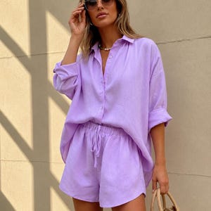 Pode incluir: Um conjunto de camisa e calções de linho lilás. A camisa de manga comprida é abotoada e os calções têm cintura com cordão. O conjunto é complementado com óculos de sol e uma bolsa de mão de vime. O conjunto é feito de tecido leve.