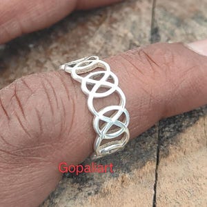 May include: A silver ring featuring a repeating openwork pattern of interconnected circles and cross shapes. The band has a polished metallic finish and a continuous design that wraps around the finger. The text Gopalart is visible in red.
