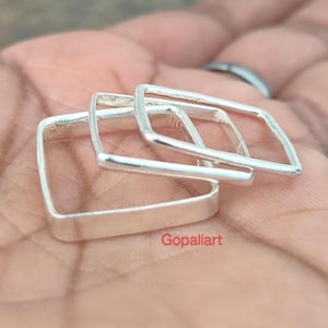 Puede incluir: Tres anillos cuadrados de plata apilados sobre una mano. Los anillos tienen un diseño minimalista con una superficie plana y pulida. Los anillos están hechos de un metal brillante y reflectante. La palabra "Gopaliart" es visible en la imagen.