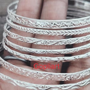 May include: A selection of silver bangles featuring detailed floral and leaf patterns. The bangles are stacked on a hand, highlighting the intricate designs. The word "Gopliart" is visible on the bottom bangle.