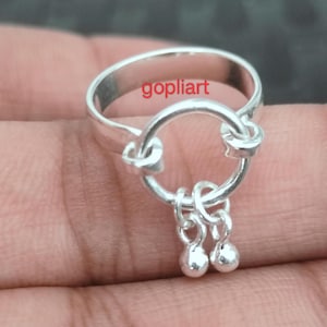 May include: A silver ring with a circular design and two dangling, small, round charms. The ring has the word "gopliart" printed on it. The ring is shown on a person's hand.