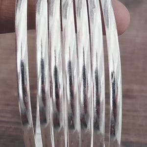 May include: A set of seven shiny, silver bangles. The smooth, rounded bracelets are stacked together, reflecting light. The word "Gopliart" is visible in red at the bottom of the image. The bangles are displayed against a blurred wooden background.
