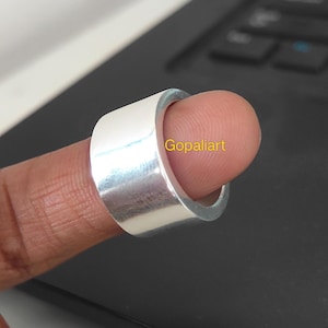 Puede incluir: Un anillo de banda ancha en tono plateado pulido con una superficie lisa y plana. El anillo presenta el texto amarillo "Gopaliart" impreso claramente en su superficie exterior. La banda tiene un ancho constante y un acabado metálico brillante.