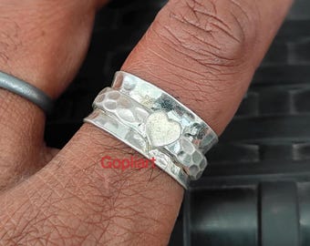 Può includere: Un anello spinner in argento con un cuore al centro. L'anello ha una texture martellata sulle fasce esterne e un cuore liscio. L'anello è indossato su un dito.