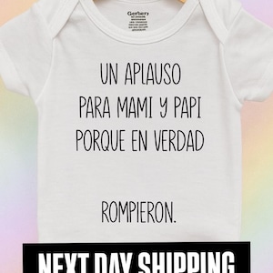 May include: White baby onesie with the text "UN APLAUSO PARA MAMI Y PAPI PORQUE EN VERDAD ROMPIERON." printed in black. The onesie is hanging on a wooden hanger.