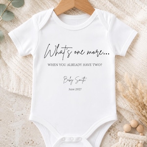 May include: A white short-sleeved baby bodysuit featuring black printed text that reads: What's one more... WHEN YOU ALREADY HAVE TWO? Baby Smith June 2027. The garment is displayed on a wooden hanger and includes a snap-button closure at the bottom.