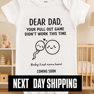 Puede incluir: Body de bebé blanco con texto negro: "DEAR DAD, YOUR PULL OUT GAME DIDN'T WORK THIS TIME" y un gráfico de espermatozoide y óvulo. Debajo: "Baby (Last name here) COMING SOON".