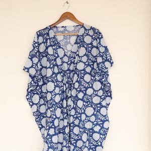 Puede incluir: Un vestido caftán con estampado floral azul y blanco colgado de una percha de madera. El vestido tiene cuello en V y cintura con cordón. El estampado floral presenta grandes flores y hojas blancas sobre un fondo azul oscuro. El caftán es de tejido ligero.