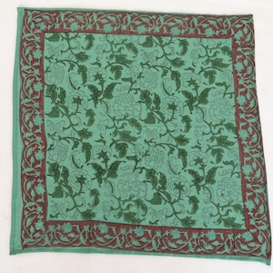 May include: A square, green textile with a floral pattern. The center features a repeating design of dark green flowers and vines. A dark brown border with a floral motif frames the edges. The textile is likely a decorative item.