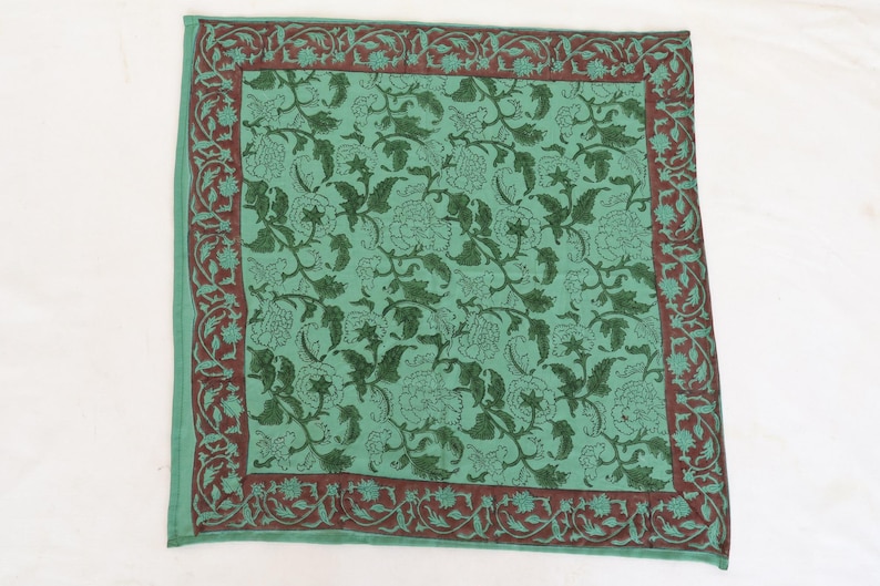 May include: A square, green textile with a floral pattern. The center features a repeating design of dark green flowers and vines. A dark brown border with a floral motif frames the edges. The textile is likely a decorative item.