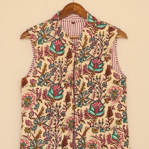 Puede incluir: Un chaleco sin mangas con un estampado floral en verde azulado, rosa y marr&oacute;n sobre un fondo crema. El chaleco tiene un forro a rayas y se abrocha con botones en la parte delantera. Est&aacute; colgado de una percha de madera.
