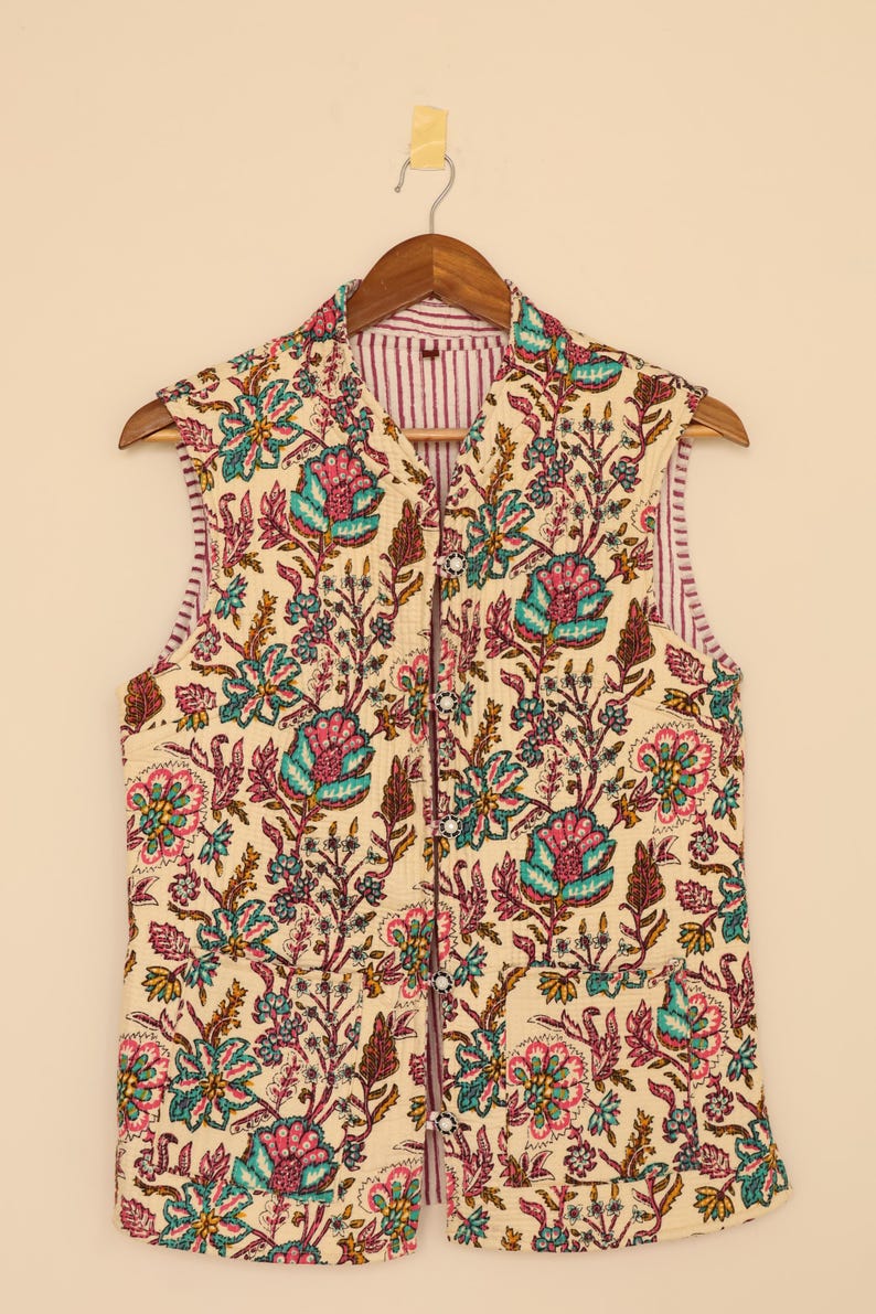 Puede incluir: Un chaleco sin mangas con un estampado floral en verde azulado, rosa y marr&oacute;n sobre un fondo crema. El chaleco tiene un forro a rayas y se abrocha con botones en la parte delantera. Est&aacute; colgado de una percha de madera.