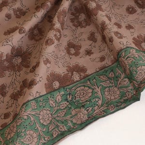 May include: A floral patterned textile with a light brown background and brown flower motifs. The border is green with floral and leaf designs. The fabric is folded, showing the pattern and texture.