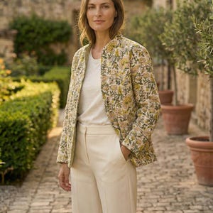 Könnte beinhalten: Eine Frau trägt eine Jacke mit floralem Muster, ein cremefarbenes Oberteil und eine Hose. Die Jacke hat ein gelbes und grünes Blumenmuster. Die Hose ist weit geschnitten. Der Hintergrund zeigt einen gepflasterten Weg und Topfpflanzen.