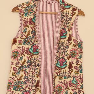 Puede incluir: Un chaleco sin mangas con estampado floral con fondo crema y un colorido estampado floral en tonos rosa, verde azulado y marr&oacute;n. El interior del chaleco presenta un estampado a rayas en blanco y burdeos. El chaleco est&aacute; colgado de una percha de madera.