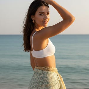 May include: A woman wearing a white ribbed bikini top and a patterned sarong on a beach. The sarong is green and yellow with a geometric design. The background is the ocean and sky.