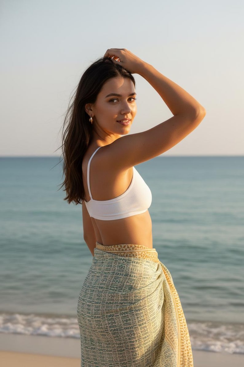 May include: A woman wearing a white ribbed bikini top and a patterned sarong on a beach. The sarong is green and yellow with a geometric design. The background is the ocean and sky.