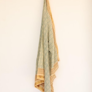 May include: A draped textile with a geometric pattern in shades of green and beige, accented with a gold border. The fabric is hanging against a white wall, showcasing its intricate design and color palette.