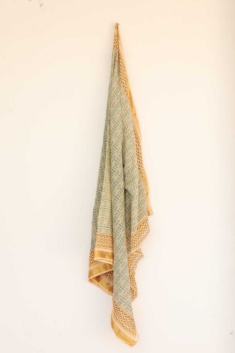 May include: A draped textile with a geometric pattern in shades of green and beige, accented with a gold border. The fabric is hanging against a white wall, showcasing its intricate design and color palette.