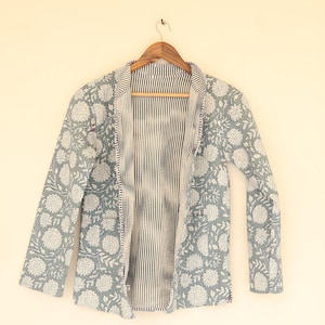 May include: A light blue quilted jacket with a floral pattern in white. The jacket has long sleeves and a striped interior. It is hanging on a wooden hanger against a cream-colored wall.