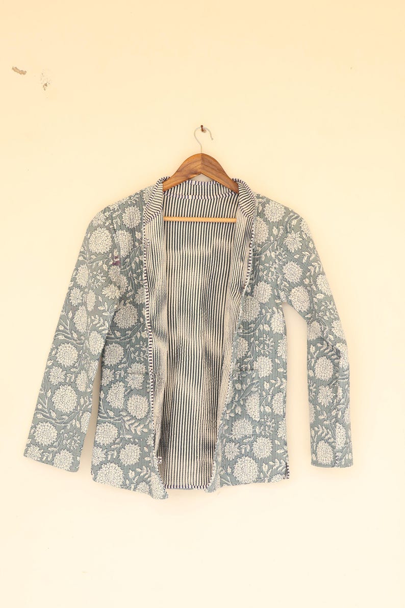 May include: A light blue quilted jacket with a floral pattern in white. The jacket has long sleeves and a striped interior. It is hanging on a wooden hanger against a cream-colored wall.