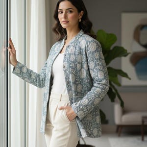 May include: A light blue jacket with a floral pattern. The jacket is worn over a white top and paired with cream-colored pants. The jacket has a button-up front and long sleeves. The outfit is completed with gold earrings.