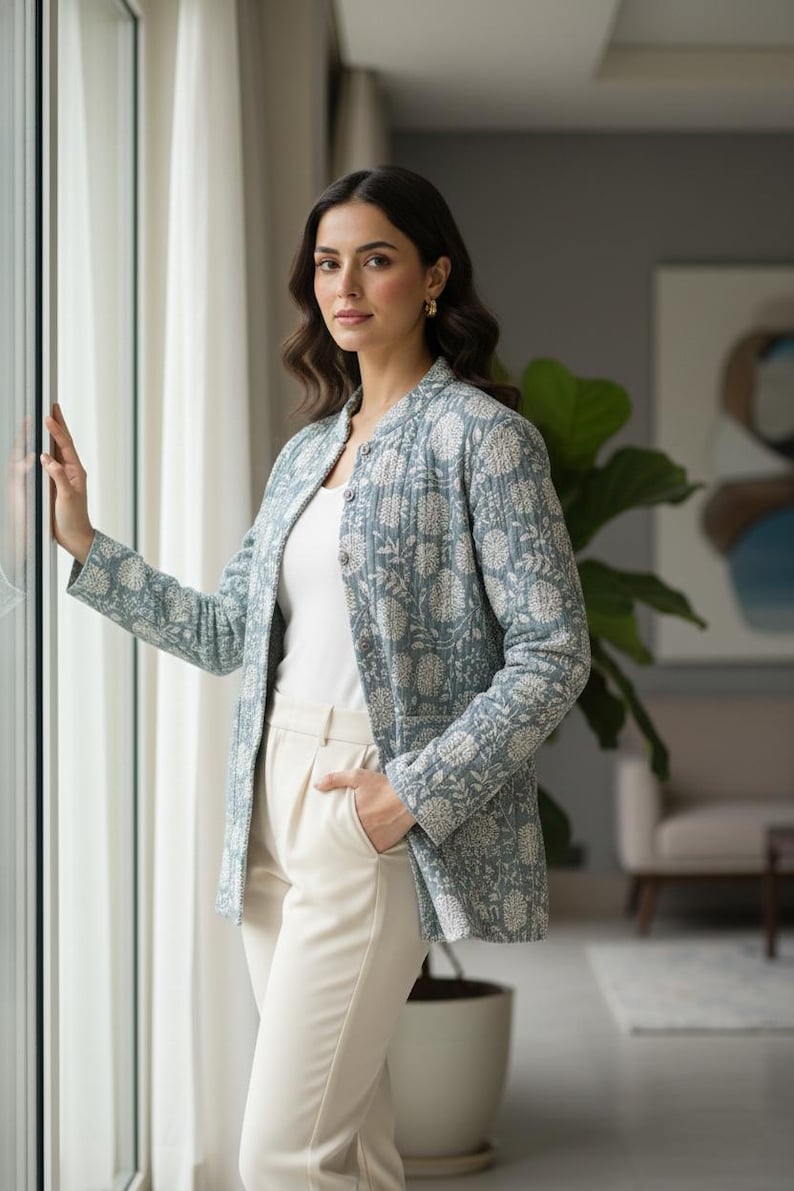 May include: A light blue jacket with a floral pattern. The jacket is worn over a white top and paired with cream-colored pants. The jacket has a button-up front and long sleeves. The outfit is completed with gold earrings.