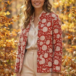 May include: A red jacket with a white floral design. The jacket has a mandarin collar and is fastened with buttons. The person is wearing a cream blouse and trousers. The background is a blurred forest scene with autumn leaves.