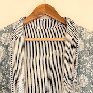 May include: A reversible jacket with a floral pattern on one side and vertical stripes on the other. The floral side features white flowers on a muted teal background. The jacket is hanging on a wooden hanger.